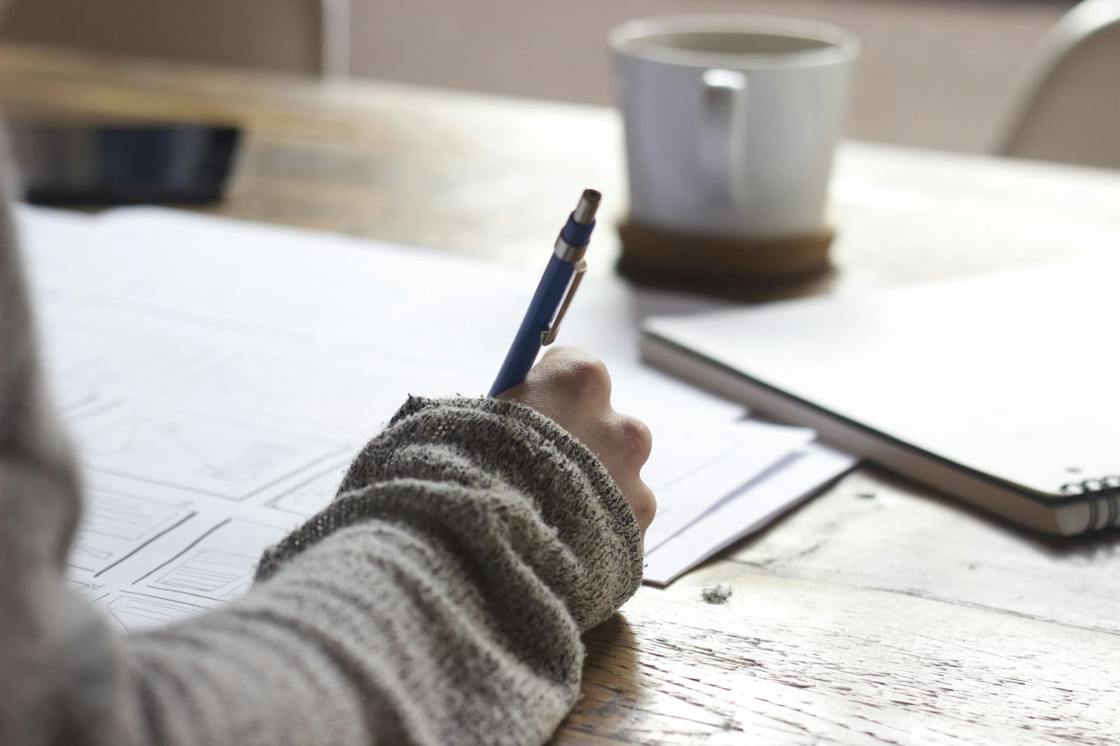 A person writing notes on paper at a wooden table, with a mug and notebook nearby.